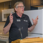 person in gray shirt speaking at a podium with out stretched hands